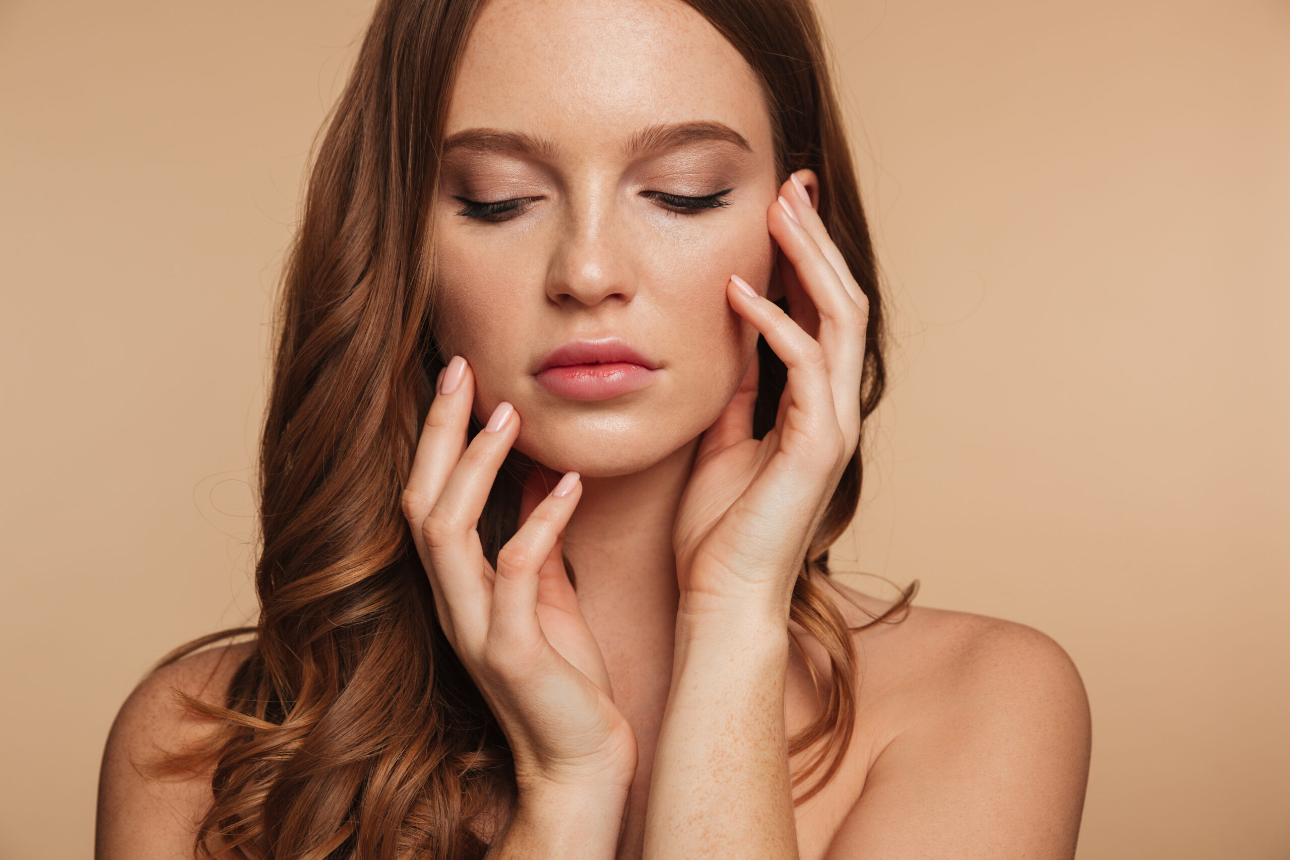 Close up Beauty portrait of sensual ginger woman with long hair posing with arms near the face over cream background